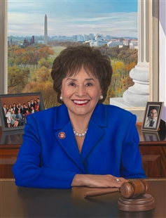 Portrait painting of Nita M. Lowey seated behind a desk with a House Member lapel pin. Her right elbow rests on the desk which has a gavel. Behind Lowey are two framed family photos. Through a window, a column base, the Washington Monument, and the National Mall are visible.