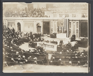 A faded black and white photograph of the crowded House Chamber with Warren G. Harding standing and speaking from the rostrum. Rows of seated Members fill the foreground and, while additional spectators stand along the upper balcony.